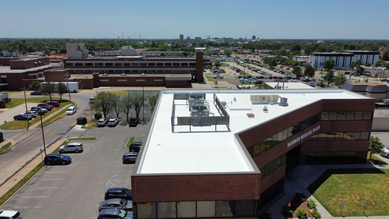 Aerial view of a commercial building with a white silicone roof coating, featuring rooftop HVAC units, surrounded by a parking lot and greenery, highlighting cost-effective roofing solutions for Oklahoma's climate.